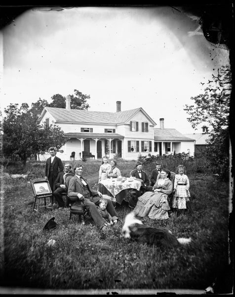 The Tollef Olsen family posed around a table in the yard with the family dog. A framed print of a long-haired retriever is placed prominently on a chair to the left. The upright-and-wing frame house has parlor furniture and a knick-knack shelf displayed on the front porch. The youngest child, Theodore Olsen, is seated at the feet of Tollef Olsen.