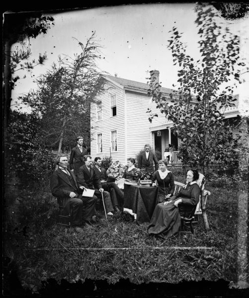 The R. Daniel Rickelsen family sitting around a table, including a small dog near the base of the table. One man is sitting and holding a cane. A lady on the right has a watch chain. Behind them is a frame house with a bird cage hanging on the porch.