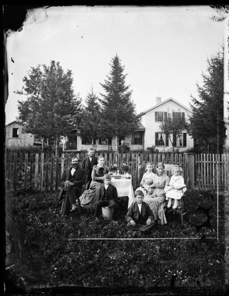 Family in yard in front of picket fence, with a farm house and a building among trees in the background. The family is gathered around a table, and a young girl is sitting in a high chair on the right. Behind the group is a man leaning on a tree in the yard behind the fence.