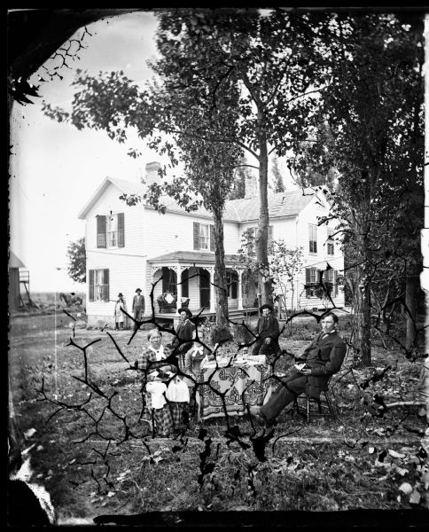 A family sits around a table in a yard. One woman holds a stereoscope in her lap. Behind them is a frame house with shutters on some of the windows, and a small loveseat on its porch. Two men are standing near the corner of the porch.