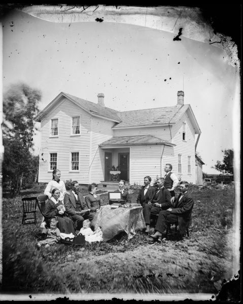 A family sits around a table on which is a wooden box with a key lock and a hinged lid, possibly with a mirror inside the lid. (A jewelry box? a music box?) The children have a doll and a doll bed. Behind the group is a frame house with double doors off the inside corner of the open porch.