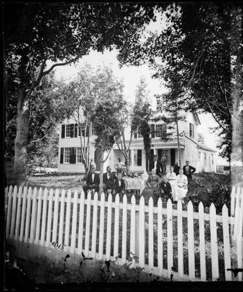 A family sits in a front yard at a table with a picket fence in front of them and a wood frame house with shutters behind. Clothes are hanging on a line on the left behind the house