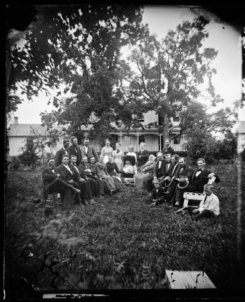 Semi-circle of group in yard with a baby carriage in the middle. A large wood frame house with carpenter's lace on its porch is behind them. The third man from the left in the back row appears to be Nels Dahl. His wife has her hand on the baby carriage. Taken at second parsonage of Herman Amberg Preus, Spring Prairie.