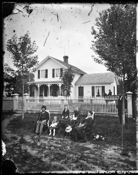A family poses in front of the picket fence before their frame house. Two little girls hold dolls in their laps. A dog sits on the grass nearby. Behind the fence near the house a boy is posed on a horse.