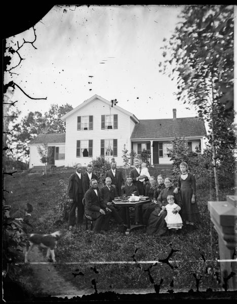 A large family is seated around a table in the yard before a large frame house with shuttered windows. A dog is in the left foreground.