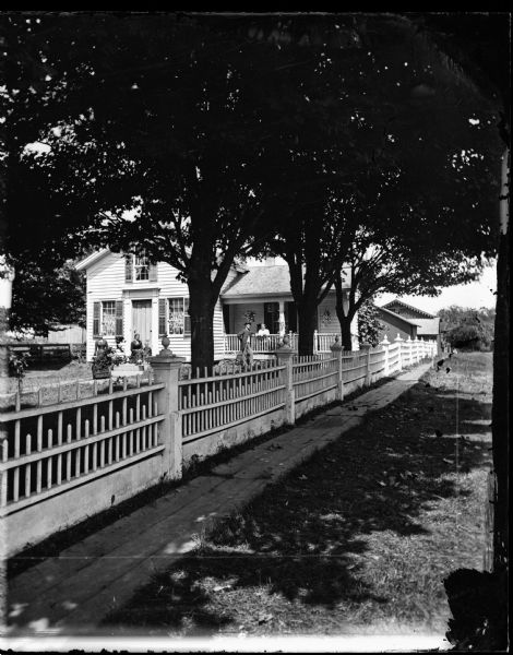 View from road down wooden sidewalk which runs adjacent to an elaborate fence along a yard, behind which women stand near the front porch of a frame house with shuttered windows. Hanging plants are on the porch.