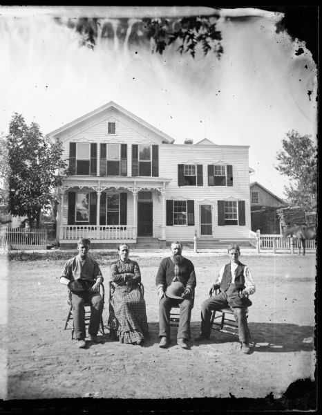 A family is seated in chairs in the middle of a street in front of a frame house with shuttered windows. Adjoining this house is what may be a commercial structure, having a false front in the style of a storefront and a hitching post in front of it.