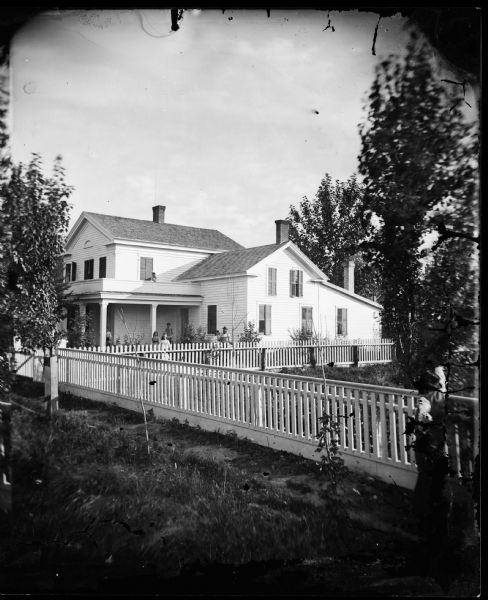 A substantial wood fence runs along and on the side of a Greek Revival house. A child is standing on the porch roof behind a railing, and other children are standing below on the porch. A man with a beard and wearing a hat is standing in the yard on the far right. He is holding a long stick or club in his hand, and the young girl standing behind him is also holding a club. Perhaps for playing croquet. Many of the wooden shutters on the house are closed.