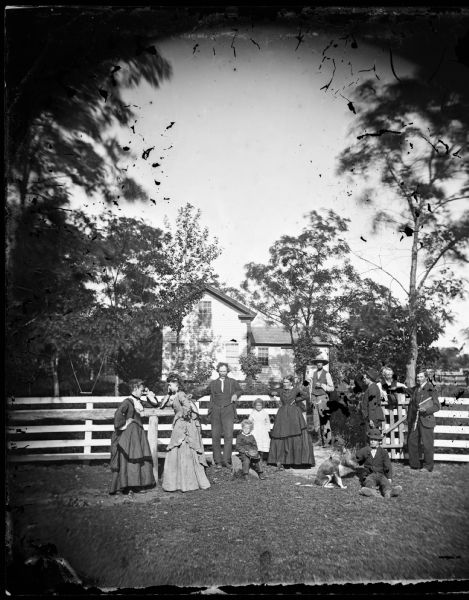 An unidentified family, thought to be Yankees, pose at a board fence in front of an upright and wing frame house. The group includes two women with poses identical to those seen in fashion prints of the day, children with the family dog, and a man at the far right holding a wooden flute and a music book in his left hand.