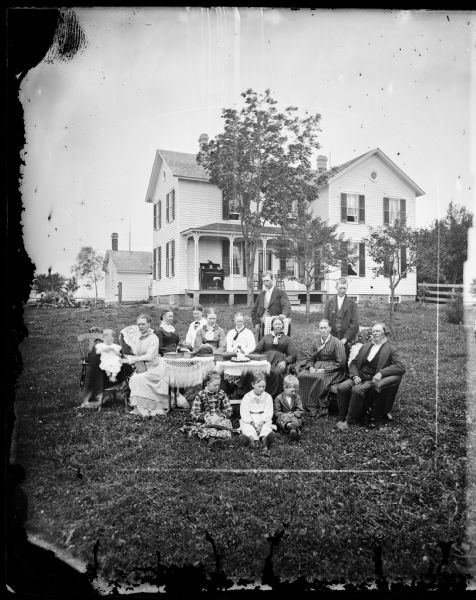 A family is in the yard around a table. A baby sits in a high chair and a man and woman are seated on a loveseat. Behind them is a frame house with an organ on its porch. A bulls-eye window is in front and on the side can be seen the stone foundation with windows. Bricks are used under the porch as supports.