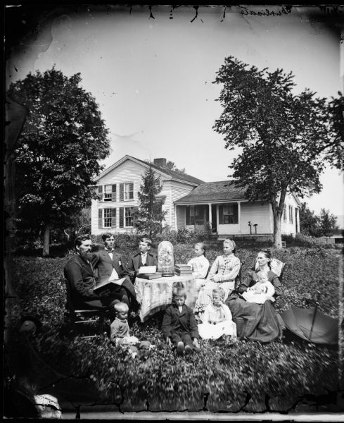 The Reverend Peter Andreas Rasmussen (1829-1898), his wife Ragnhild Holland Rasmussen, and their eight children sit around a table in the yard of their house. The table is burdened with large books and a large glass dome containing a floral arrangement. Behind them is an upright and wing frame house.