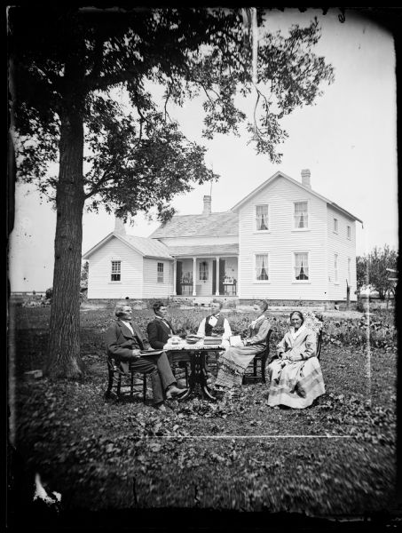 The Ashburn family sits around a table in their yard. Behind them is a frame house with plants on its porch, curtains visible in its windows, and a pump by its side. Left to right: Jacob Larsen, Ole Ashburn, Daniel Skare (brother of Ole Ashburn), Julia Larsen Ashburn (daughter of Jacob and wife of Ole) and Anna Larsen (stepmother of Julia and second wife of Jacob). Lived NW 1/4 or SW 1/4 Section 36, Town of Clinton, Rock County, 1/2 mile north of the Illinois border in Jefferson Prairie area.<br>In this photograph, Daniel Skare is visiting U.S. from Norway. He didn't immigrate until 1880. Then he attended Beloit Seminary in Iowa and became a minister. Ole and Julia were married about this time, late June, 1877.</br>