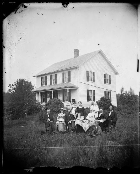 A family is gathered around table for coffee with a frame house behind that has a sewing machine on its porch, and a lightning rod on the roof. A framed picture is hanging on the porch.