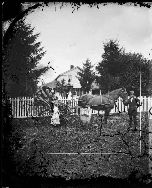 A man with an impressive beard, and a woman, are sitting together in a carriage with a collapsible top. A young girl is standing by the carriage while holding the man's hand. On the right is a man standing and holding the head of the horse pulling the carriage. Behind them is a picket fence, and a woman is standing on the porch of a frame house that has latticework on all sides of the porch.