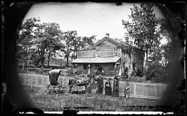 A carriage and a family stand in front of a picket fence in front of an unpainted Greek Revival frame house. Attached to the house is a storefront. Although unidentified, the fact that the photograph is part of the collection of Andrea Dahl, suggests that the location is somewhere in Dane County.