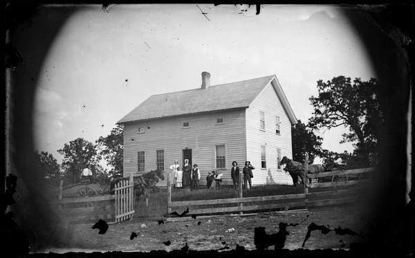 View uphill towards a family standing in a yard, some in wagons, behind a wood fence and picket gate. Behind them is a simple frame house with small windows on the second story under the eves.