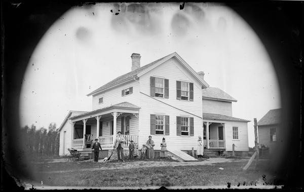 A family is posing at the side of a house. A man and boy are holding rifles, and a dog is lying on the ground between the. There is a baby sitting in a high chair near the woman in the center. The house has latticework on both the front and back porches, and some carpenter's lace. There are half windows on the second story under the eaves. There is a slanted cellar door (bulkhead) that extends into the yard. Frame outbuildings are in the background.