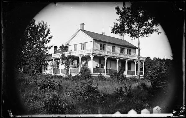 View across yard with tall plants and grass towards a man and woman sitting on a porch behind a latticework railing. The porch has carpenter's lace and a parapet roof and covers two sides of the house. Text on back of stereograph print reads: "R.E. Davis Residence, Davis is on porch - became Senator".