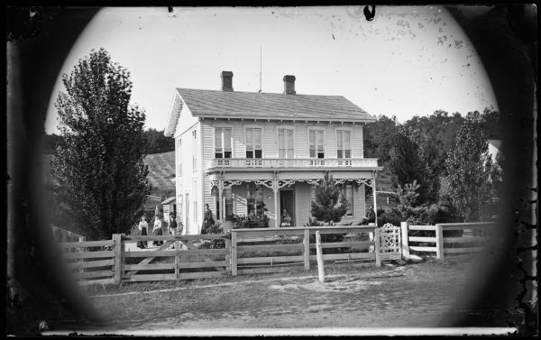 Exterior view of the Kingsley house with family in front. It has tall and narrow double-hung windows that are grouped in twos and etched. Carpenter's lace adorns the porch and an elaborate bracket-style gate adorns an otherwise plain fence in the foreground. There is a hill behind the house. Middleton (Clinton's Addition).