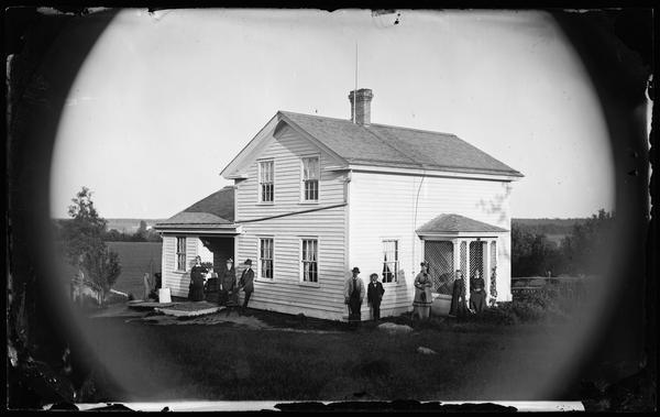 Slightly elevated view of a family standing near a small and neat rural frame house. The women are wearing hats. One man is leaning against the house with one foot on a chair. The frame house has a small front porch with latticework and a pump on the side. A small frame shed and fields are in the background.
