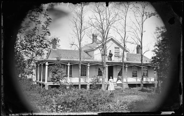 An unusual long and low frame house with a small central second story. The porch wraps around two sides and has no railing. There are some Gothic Revival decorative elements. Two women pose in front of the house and a boy sits in a chair on the porch roof with a fiddle.
