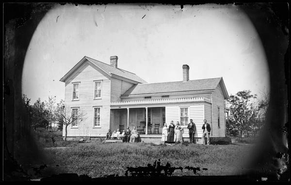 A family of eleven is sitting on the porch steps and standing in the yard alongside a wood frame house with elegant trim above its windows, and along top front of the porch roof. Four chairs sit empty on the porch.