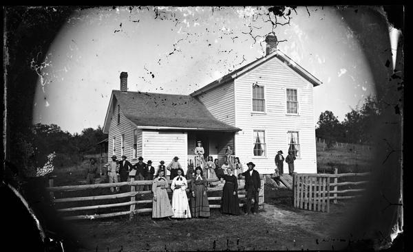 Extended family group of twenty-two people, some in work clothes, some elegantly dressed, stand in a yard and on the porch steps of an upright and wing frame house. A crude fence is in the foreground, and the cellar entrance can be seen on the right.