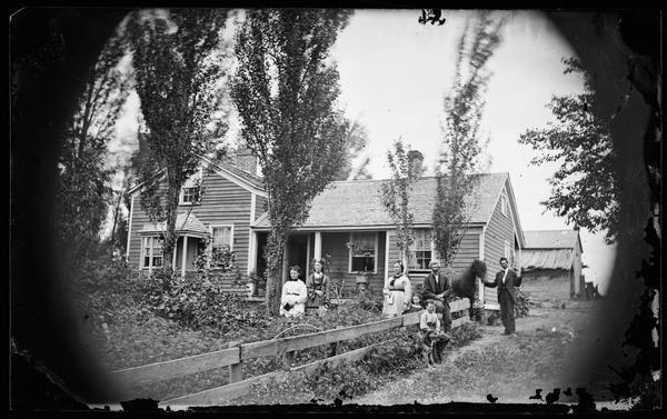 A family of seven standing in a yard near a board fence in front of their frame house. A dog is sitting near the boy in the foreground. A man standing on the far right is holding a black horse. An outbuilding is behind the house.