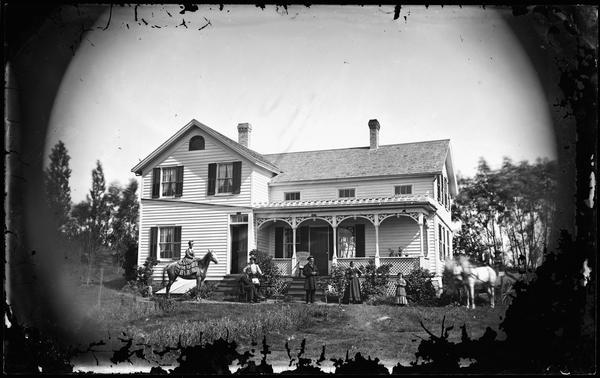 A family poses in a yard with one elegantly dressed young woman on horseback. The frame house has an unusually shaped attic story fan window. The porch is adorned with latticework and its brackets and parapet roof have attractive trim. A "Madison Mutual" sign is above the door. An exterior entrance to the cellar is to the left of the porch.