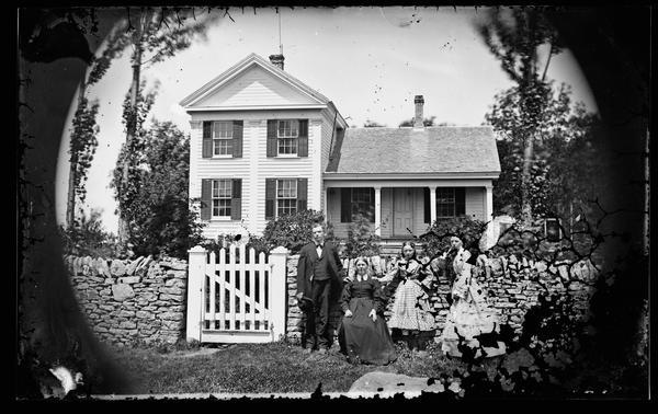 A well-dressed family of four is posed in front of a stone fence with a picket gate. Behind them is a Greek Revival frame house with shuttered windows.