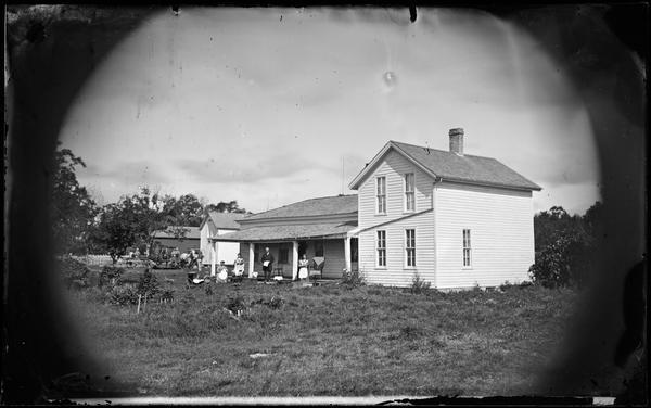 A family is posing in the yard of a farmhouse near the porch. A child is sitting in a baby buggy. Farm buildings, farm implements, wagons and people are in the background on the left.