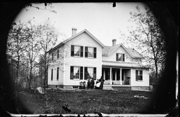 A family of seven, including a babe in arms are in front of a frame house with a stone foundation and latticework under the porch. A tree stump is in the left foreground and a rain barrel is at the right corner of the house.