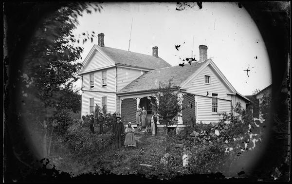 A family of nine, and a dog, are posing standing in the yard of an upright and wing house with a porch nearly completely walled-in by latticework.