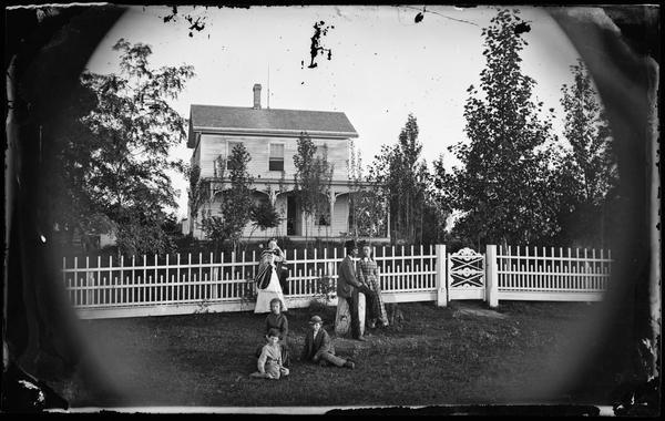 A family of six are posing in front of a fancy picket fence with an ornate Norwegian folk-style gate. The two-story frame house has a front porch decorated with carpenter's lace. A man is sitting on a tree stump mounting block wearing a top hat, and a woman on the left leaning on the fence is wearing a Norwegian-style striped shawl. Three children are sitting on the ground.