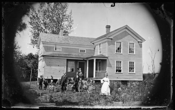 A family of eleven in the yard and on the porch, including an elderly woman holding a baby. The house is Greek Revival with a small porch and small windows in upper story in front.