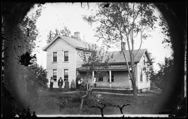 Family lined up in the yard and on the porch of an upright and wing frame house.
