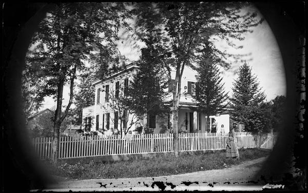Four children and a couple stand in a yard behind a picket fence and before a Greek Revival frame house with shutters.