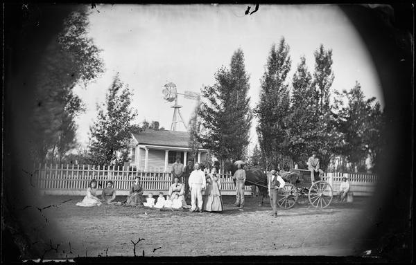 Sixteen people, including four small children dressed in white, are posed in front of a picket fence standing before a small frame house. A windmill and horse and wagon are also prominent in the scene. The windmill is a patented design by W.D. Nichols (patents of January 1, 1867 and April 19, 1870) and was manufactured by the Challenge Mill Company of Batavia, Illinois.