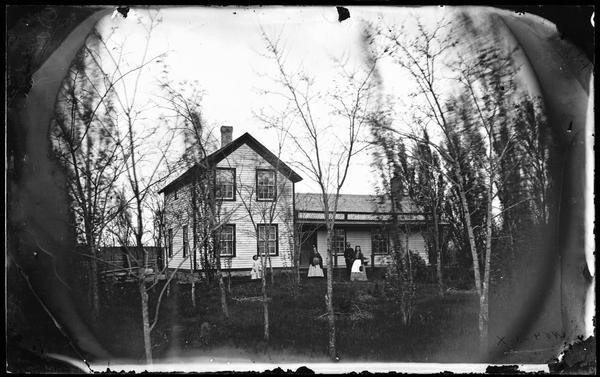 A couple and two girls stand among small trees in front of an upright and wing frame house with latticework above the porch.