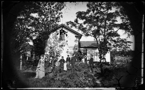 Family posing in a wooded yard. One man holds a wooden flute. Behind them is a Greek Revival house with a Madison Mutual insurance sign over the door.