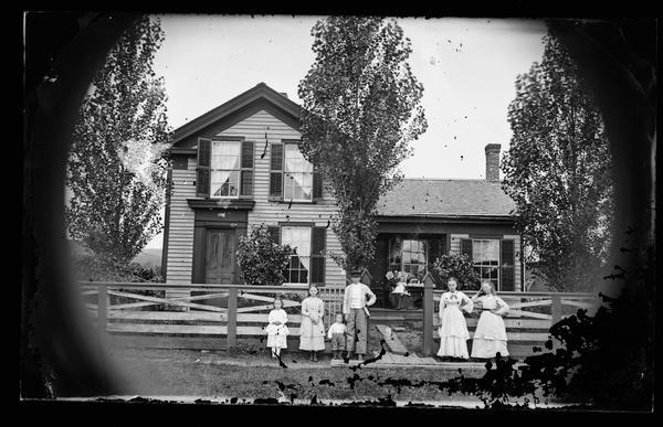 Six children standing in front of a wooden fence that has posts and a picket fence gate. A cat is sitting on the wooden walk behind them and their mother is sitting on the porch with a baby. The Greek Revival house has the Norwegian-American Hekla Fire Insurance Company sign over its door. The linear organization used here was one adopted by Dahl early in his career. Said to be the Al Meyer residence, 7400 block of Elmwood, in Middleton.