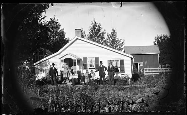 A family of seven is posed around a table in the yard of a very modest house with a latticework porch. The men wear hats and a woman holds a baby. The house's chimney is made of brick and pipe. A fence and other frame buildings are behind them.