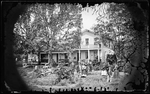 The Herman Amberg Preus family and visitors, all wearing hats, are playing croquet in their yard. One child is on a rocking horse and another child is by a baby carriage. Rustic wooden benches are under the trees near the porch. The frame house behind them has latticework on its porch. Walworth Co.