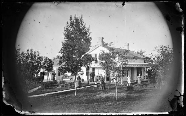 Four women in yard, one holding a doll with a woman sitting on the porch behind frame house. There is a man holding a shovel and two children are standing on the porch roof. The house has woodwork trim at porch top, and potted plants on side porch.