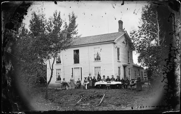 Fifteen adults are seated around table being served coffee in the yard. One woman sits at the spinning wheel and another at a sewing machine. The Greek Revival frame house has an open second-story door with two children standing in it.