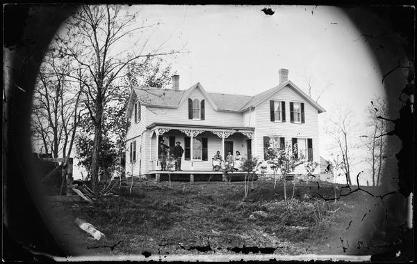 Two men standing and three women sitting on porch that has carpenter's lace trim; frame house with gable and window and latticework at bottom of porch. A dormer above the porch has a tablet style window with rounded shutters. Newly planted trees are in the yard.
