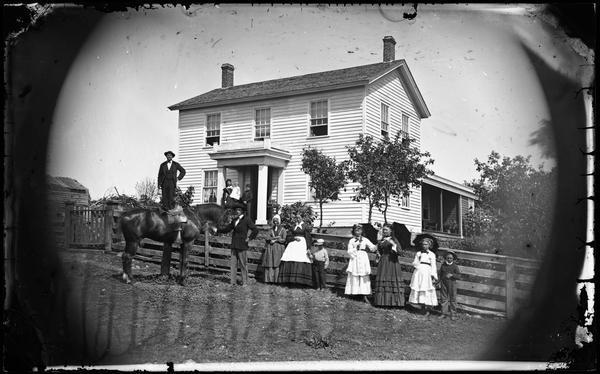 A family poses in front of a fence, including women with parasols, a man with a horse, a man standing on a fence and a woman and two children on the porch of the frame house in background. One woman is wearing a hoop skirted dress.