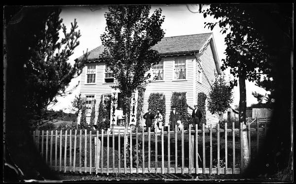 A wooden fence with widely spaced pickets is in the foreground. Behind it is a family in the yard of a frame house that has vines covering its windows, nice trim along the roof edge and similar porch trim including latticework. An animal, possibly a toy, is on the fence.