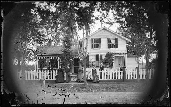 Family in front of picket fence and wood walkway, which encloses an upright and wing frame house with elaborate carpenter's lace and latticework on the porch and screen doors.