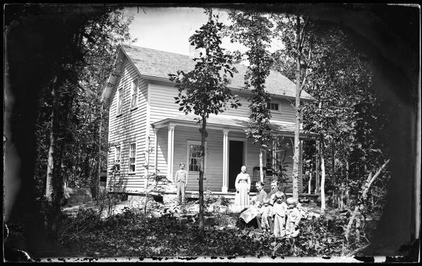 A mother, father, four children and two babies in a yard with a frame house behind, in a heavily wooded area.
